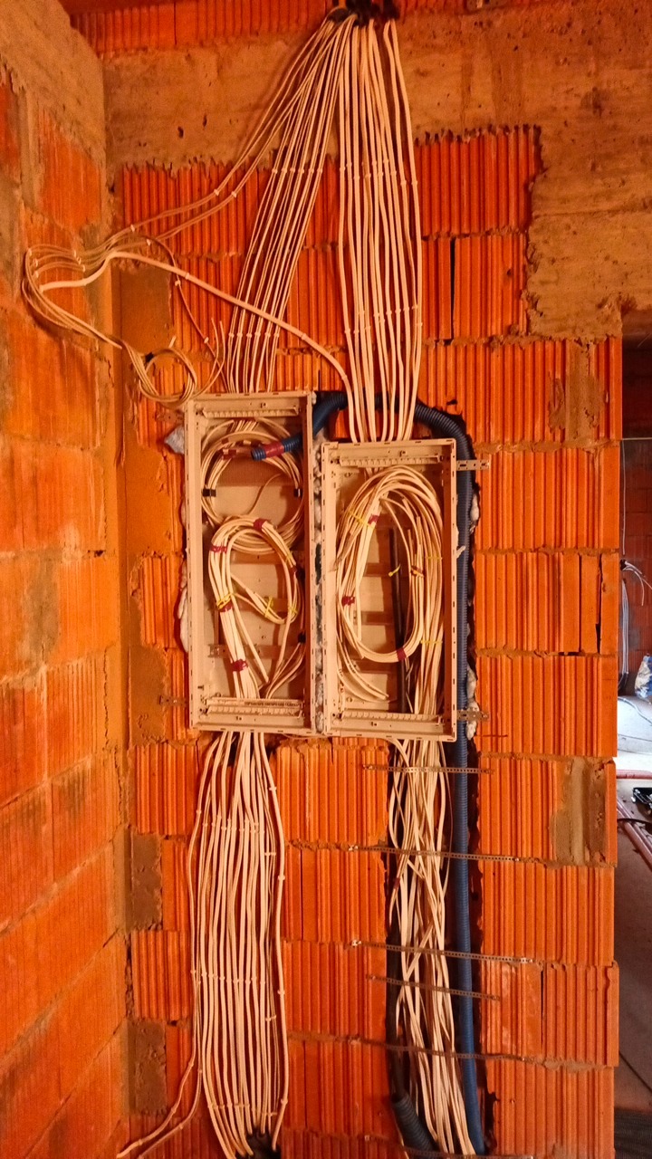 Dwa połączone rozdzielacze elektryczne z licznymi przewodami na ceglanej ścianie. Widoczne kable w peszlach i luźne przewody. Instalacja w trakcie montażu.