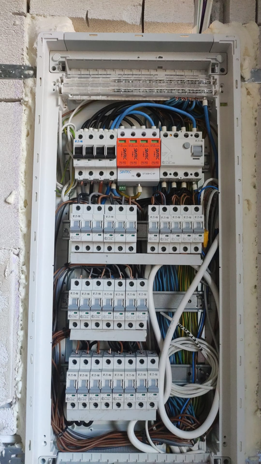 Rozdzielnica elektryczna w białej obudowie, widoczne zabezpieczenia SIMTEC i EATON, kable w różnych kolorach. Instalacja w surowym stanie, ściana z białych bloczków.