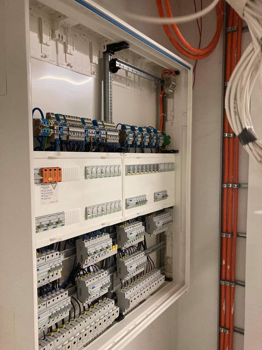 Rozdzielnia elektryczna w białej obudowie z licznymi zabezpieczeniami, przewodami i elementami montażowymi. Widoczne kable w peszlach na ścianie.