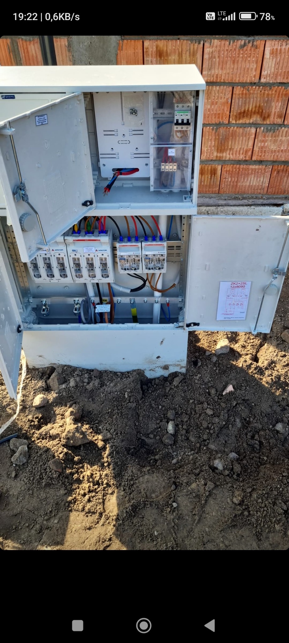 Otwarta skrzynka elektryczna z bezpiecznikami i przewodami na tle ceglanego muru, umieszczona na ziemi. Widoczne elementy instalacji i schemat.