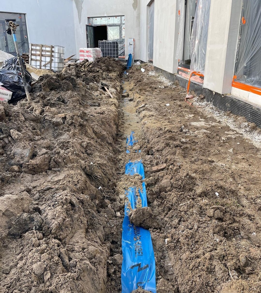 Wykop z ułożonymi niebieskimi rurami w folii ochronnej. W tle budynek w trakcie budowy z widocznymi oknami i drzwiami. Prace ziemne.