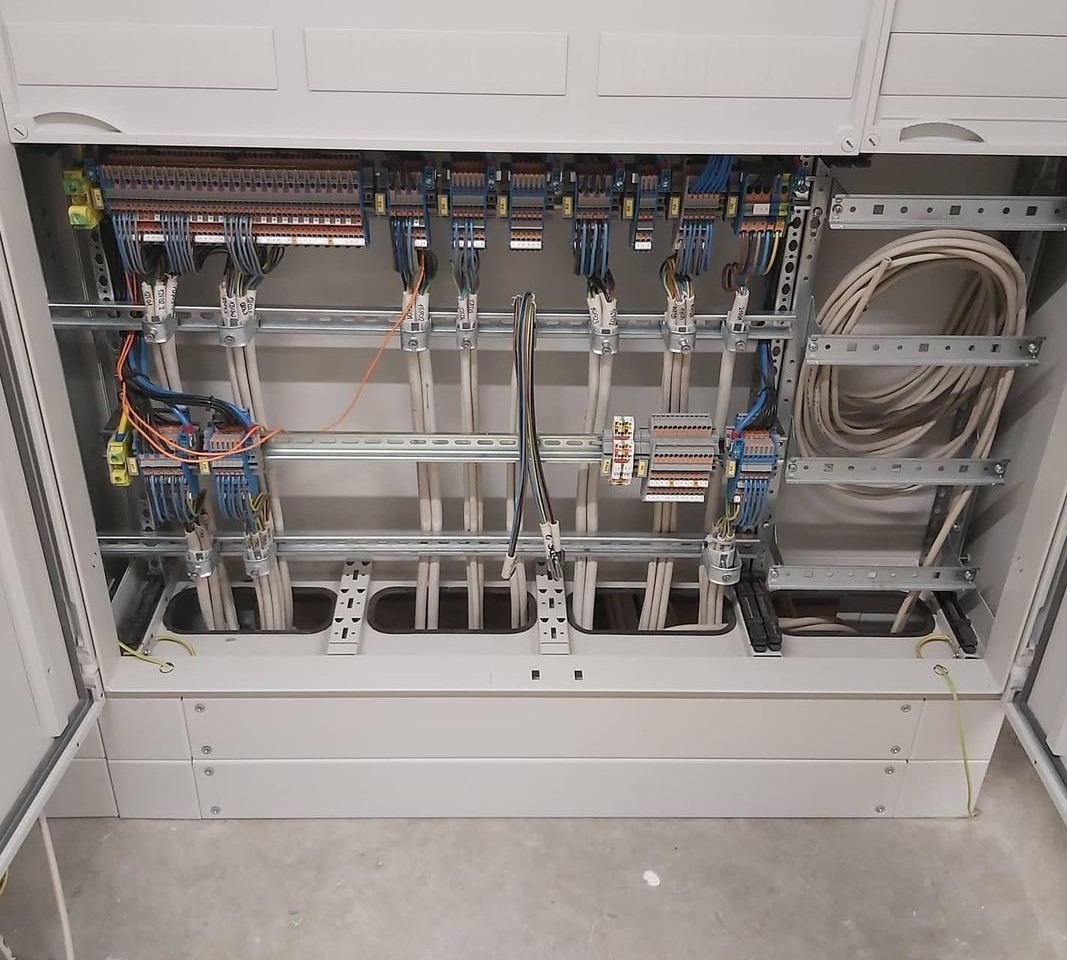 Rozdzielnica elektryczna w szarej obudowie z widocznymi kablami, listwami zaciskowymi i elementami montażowymi. Widok z góry na otwartą skrzynkę.