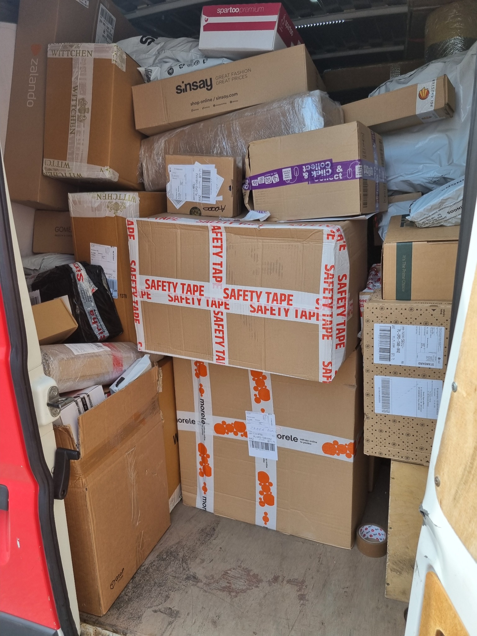 Wnętrze busa wypełnione kartonami różnych marek (Wittchen, Zalando, Sinsay, Morele) i rozmiarów, zabezpieczonymi taśmami. Widoczne etykiety adresowe.