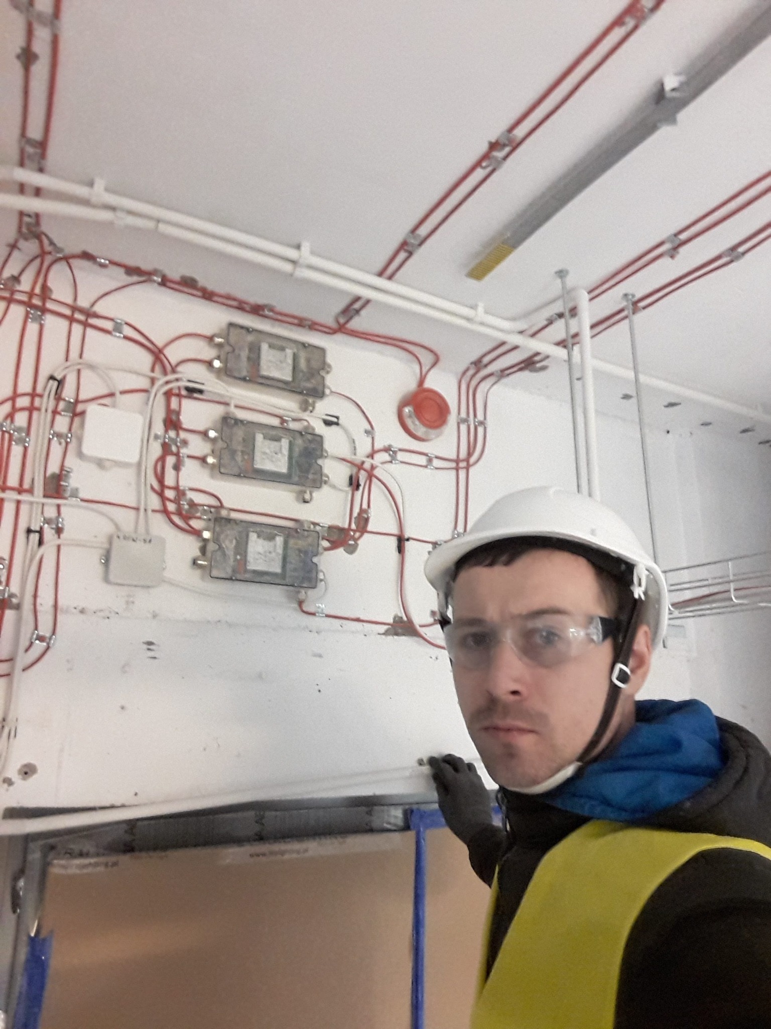 Instalator w kasku i okularach ochronnych prezentuje rozbudowaną instalację elektryczną z przewodami i rozgałęźnikami na białej ścianie wewnątrz budynku.