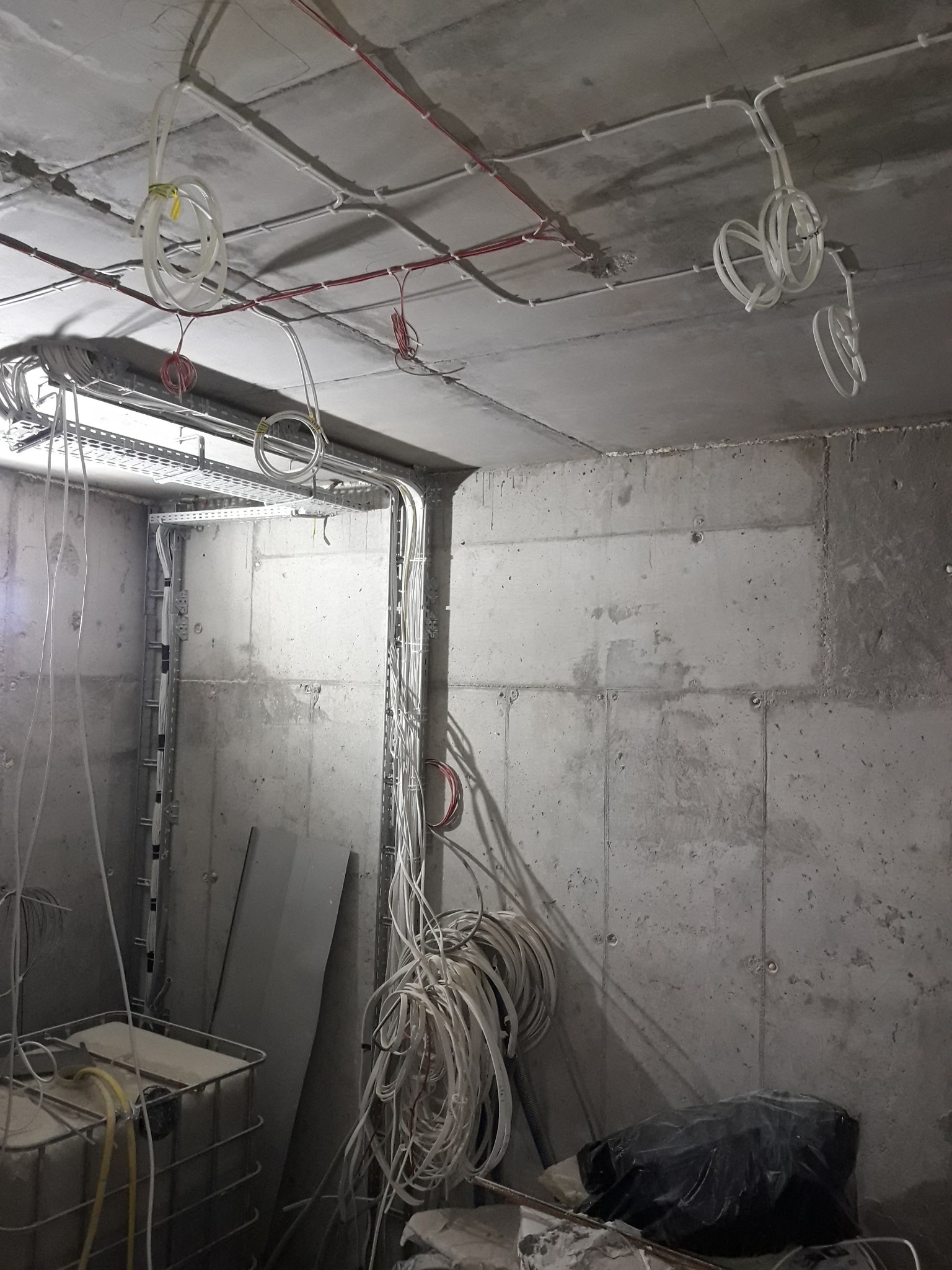 Surowy betonowy pokój w trakcie instalacji elektrycznej: widoczne przewody, kable i metalowe korytka na suficie i ścianach. Stan surowy, bez wykończenia.