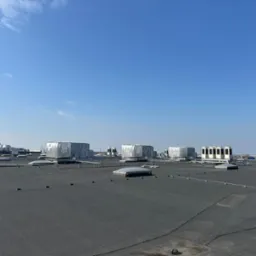 Rozległy dach płaski z licznymi urządzeniami wentylacyjnymi i klimatyzacyjnymi w srebrnych obudowach, antenami i świetlikami, na tle błękitnego nieba.