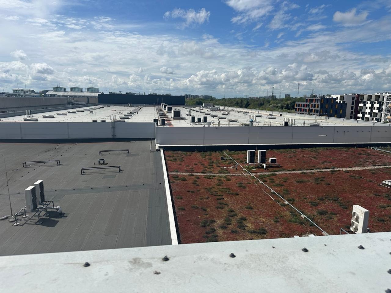 Rozległy dach budynku komercyjnego z licznymi klimatyzatorami, wentylacją i fragmentem zielonego dachu. Widok z góry, w tle błękitne niebo z chmurami.