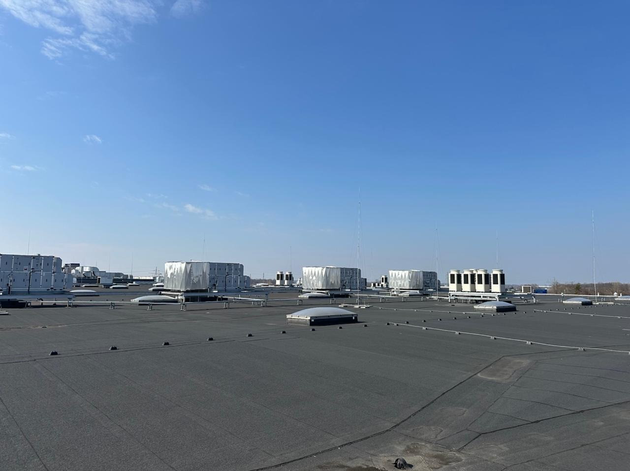 Rozległy dach płaski z licznymi urządzeniami wentylacyjnymi i klimatyzacyjnymi w srebrnych obudowach, antenami i świetlikami, na tle błękitnego nieba.