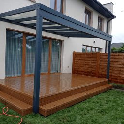 Dach i Dech Zapiera Norbert Staszewski - Nowoczesna pergola z szarą konstrukcją i drewnianym tarasem na trawie, przylegająca do jasnego domu z dużymi oknami. Widoczny fragment drewnianego ogrodzenia.