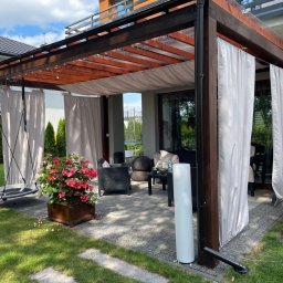 Dach i Dech Zapiera Norbert Staszewski - Nowoczesna pergola z ciemnego drewna z białymi zasłonami, huśtawką i kwiatami w donicy. Widoczny fragment domu z balkonem i zadbany trawnik.