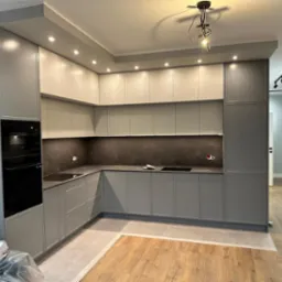 Nowoczesna kuchnia w odcieniach szarości i bieli, z drewnianą podłogą i subtelnym oświetleniem punktowym. Minimalistyczny design z zabudowanym sprzętem AGD.