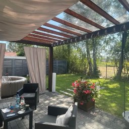 Dach i Dech Zapiera Norbert Staszewski - Zadaszona pergola z drewnianą konstrukcją i zasłonami, otoczona zielenią. Na tarasie meble ogrodowe i jacuzzi. Widoczne elementy dekoracyjne i oświetlenie.