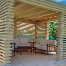 Dach i Dech Zapiera Norbert Staszewski - Drewniana pergola z ażurowymi ścianami, wyposażona w sofę i stolik kawowy, tworząca zaciszną strefę relaksu na tarasie. Widok na zieleń w tle.