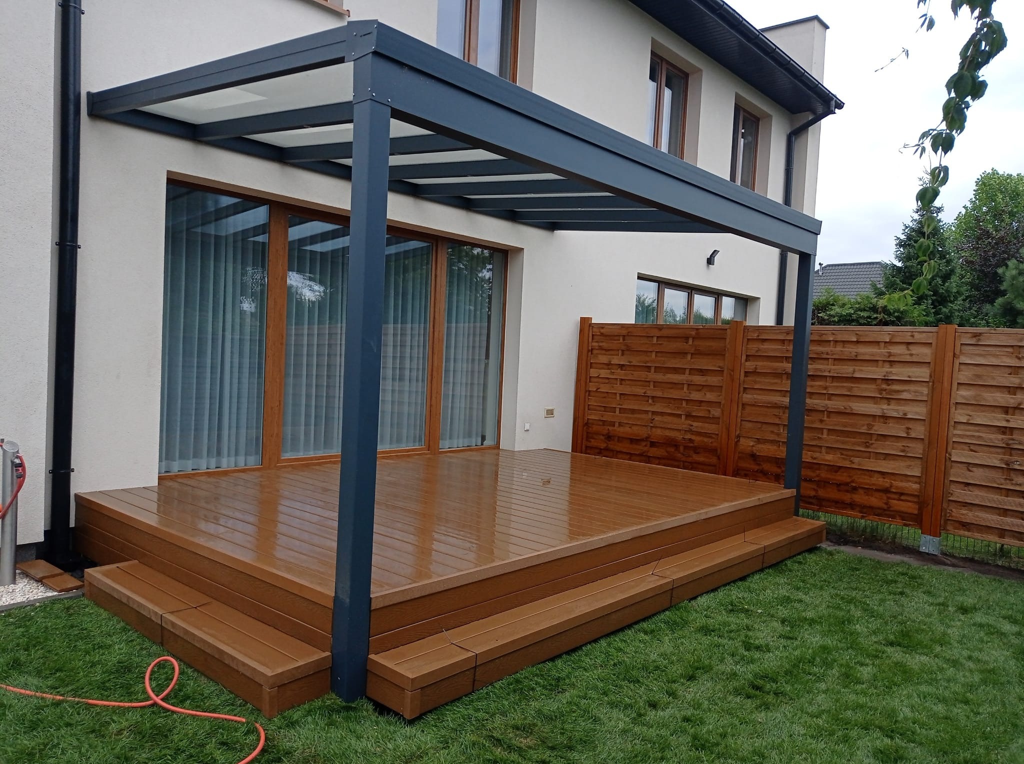 Nowoczesna pergola z szarą konstrukcją i drewnianym tarasem na trawie, przylegająca do jasnego domu z dużymi oknami. Widoczny fragment drewnianego ogrodzenia.