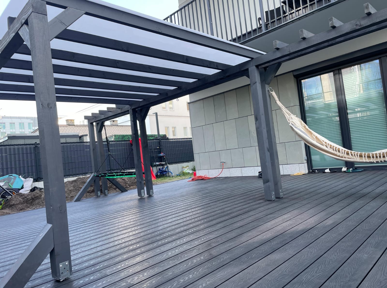 Nowoczesny taras kompozytowy w antracytowym odcieniu z drewnianą pergolą i hamakiem, przylegający do budynku z dużymi oknami. Widoczne elementy montażowe.