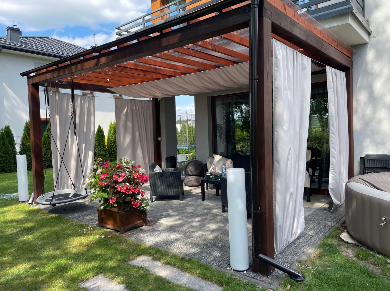 Nowoczesna pergola z ciemnego drewna z białymi zasłonami, huśtawką i kwiatami w donicy. Widoczny fragment domu z balkonem i zadbany trawnik.