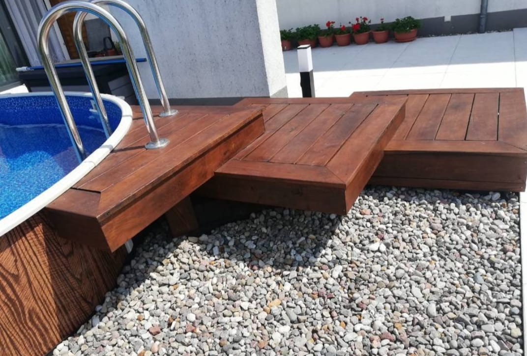 Drewniany taras przy basenie z drabinką, wykończony drobnymi kamieniami. Ciemne drewno, nowoczesny design, rośliny w donicach w tle.