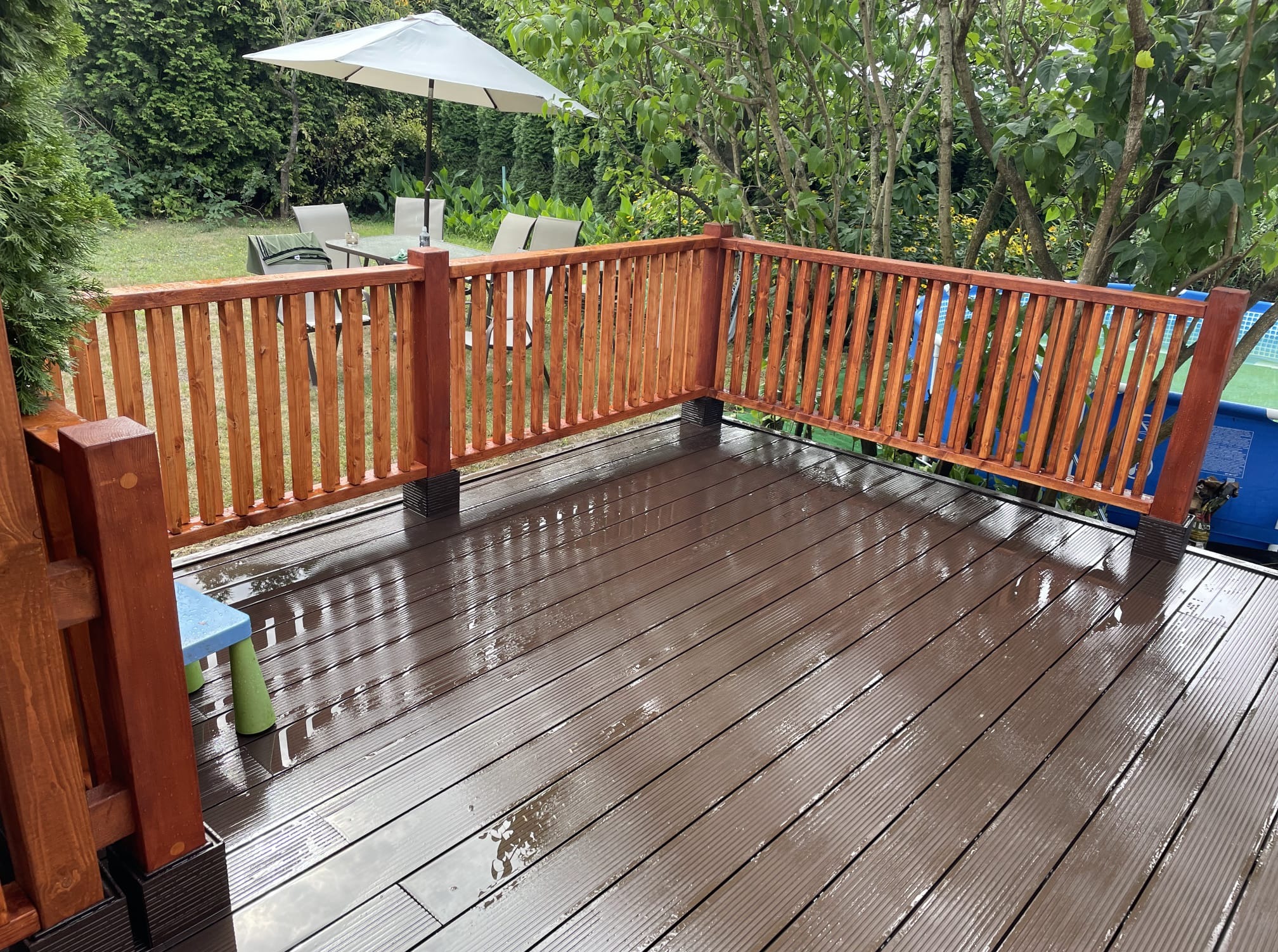 Wilgotny taras kompozytowy z drewnianą balustradą w ogrodzie, widok na meble ogrodowe i basen. Po deszczu. Deski ułożone równolegle.