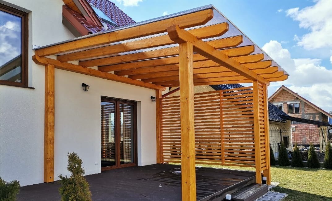 Drewniana pergola z przezroczystym zadaszeniem przylegająca do białego domu. Widoczne ażurowe panele boczne i fragment tarasu. W tle dom sąsiada i błękitne niebo.