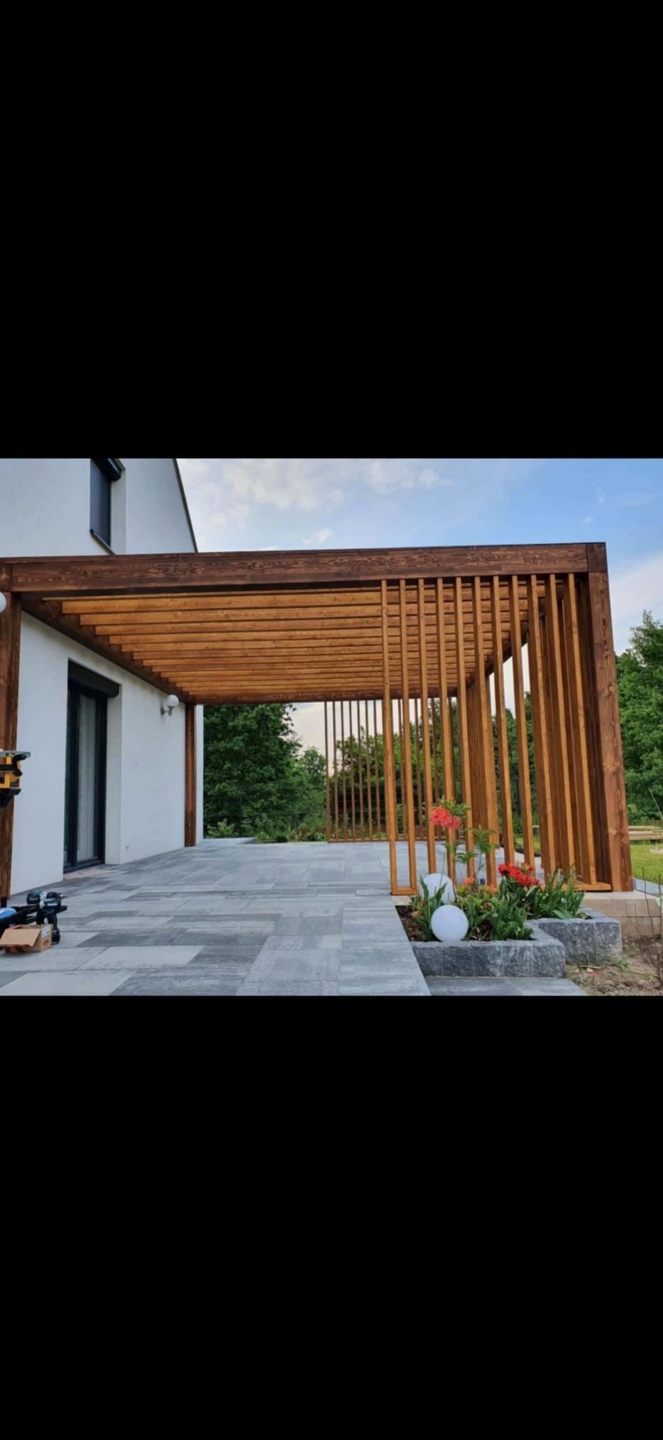 Nowoczesna pergola z drewna w kolorze orzecha, przylegająca do białego domu z szarą kostką brukową, z roślinami w kamiennej rabacie. Oświetlenie zewnętrzne.