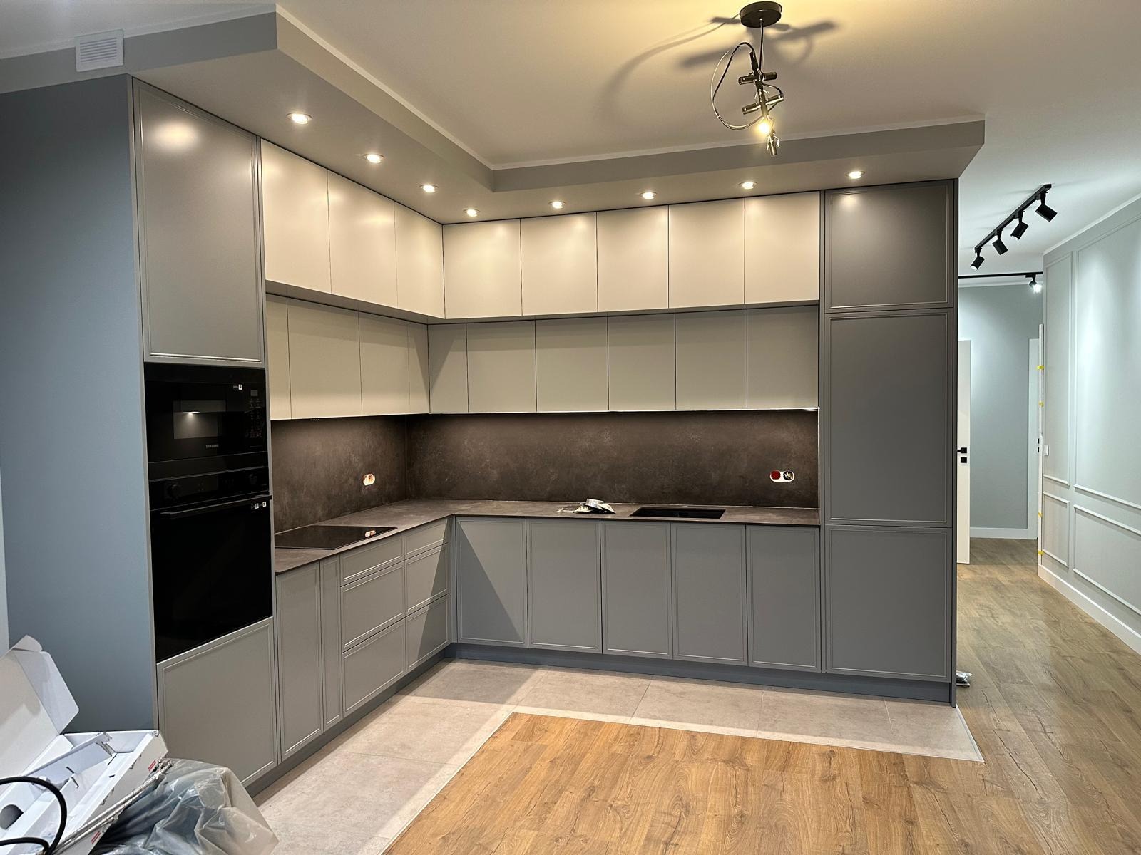 Nowoczesna kuchnia w odcieniach szarości i bieli, z drewnianą podłogą i subtelnym oświetleniem punktowym. Minimalistyczny design z zabudowanym sprzętem AGD.