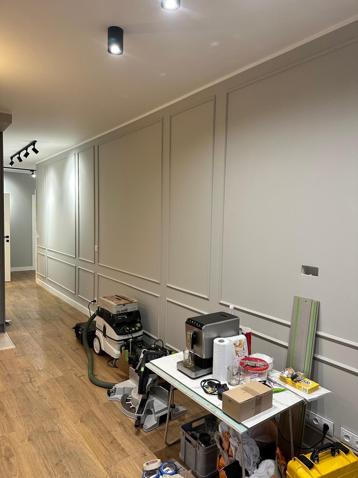 Korytarz z panelami ściennymi w stylu angielskim, drewnianą podłogą i narzędziami w trakcie wykańczania. Widoczny odkurzacz przemysłowy i stół z akcesoriami.