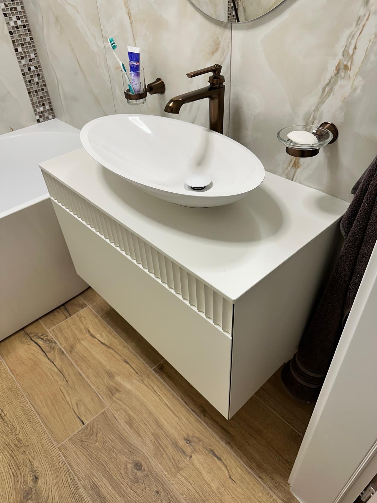 Nowoczesna łazienka: biała szafka z umywalką nablatową o owalnym kształcie, brązowa armatura i marmurowe płytki. Elementy wystroju: mydelniczka, szczoteczka do zębów.