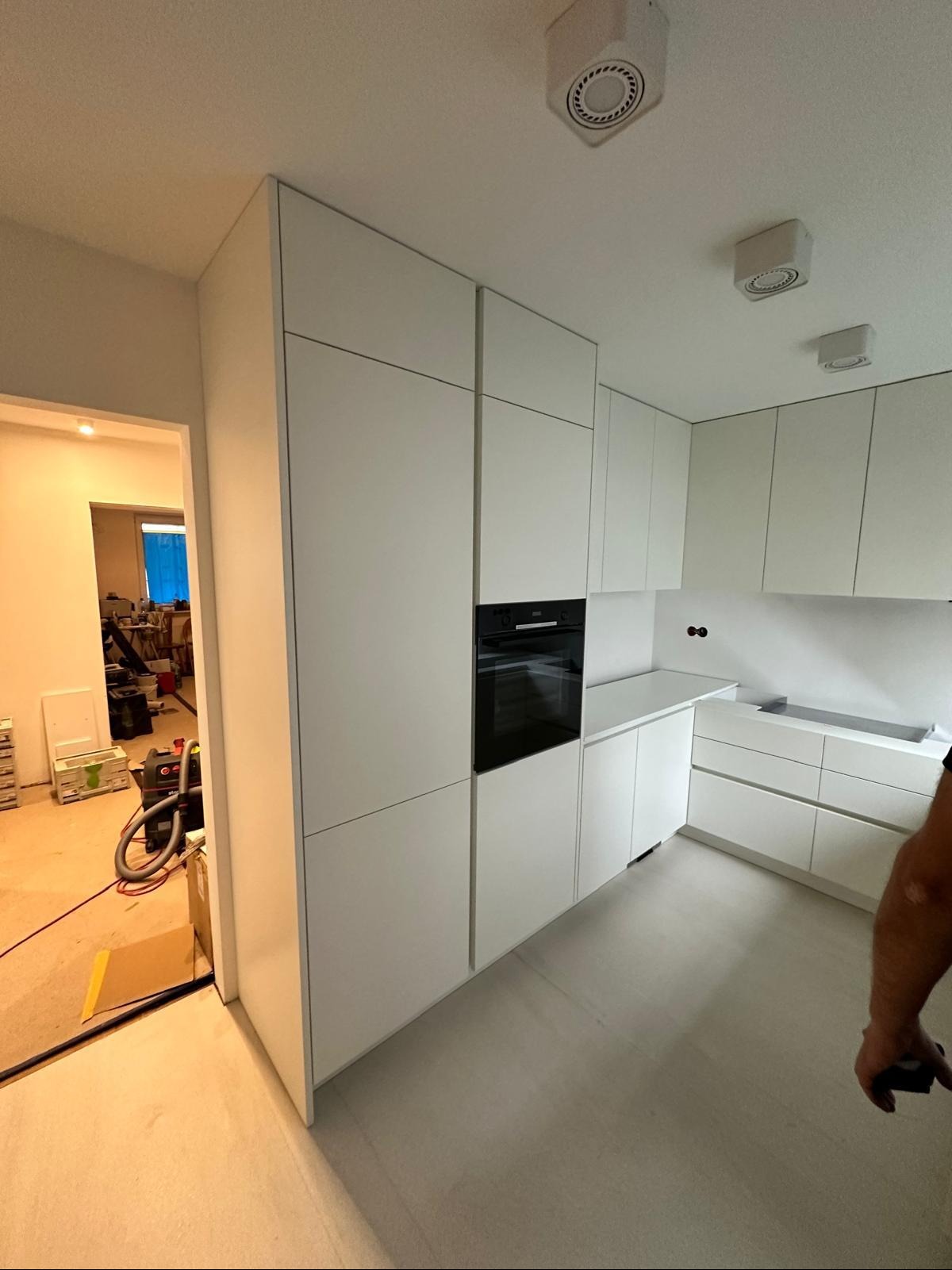 Minimalistyczna, biała zabudowa kuchenna z piekarnikiem w wysokiej szafce, widok z boku, z otwartym przejściem do pomieszczenia w trakcie prac wykończeniowych.