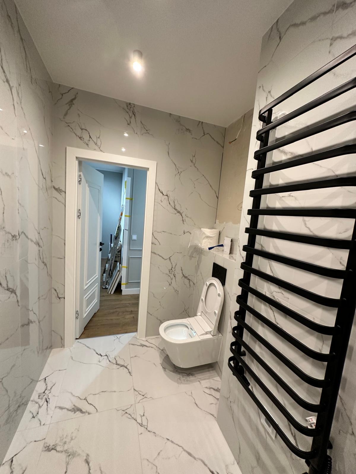 Wykończona łazienka z marmurowymi płytkami, wiszącą toaletą i czarnym grzejnikiem drabinkowym. W tle widoczny otwarty pokój z drabiną malarską.