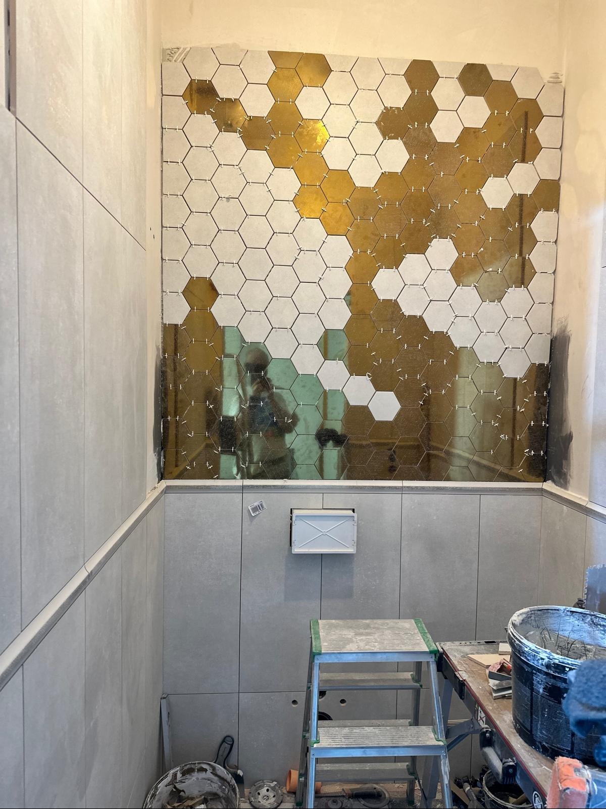 Układanie heksagonalnych płytek w łazience, częściowo wykończona ściana z białych i złotych płytek. Widoczne narzędzia i drabina, etap prac wykończeniowych.