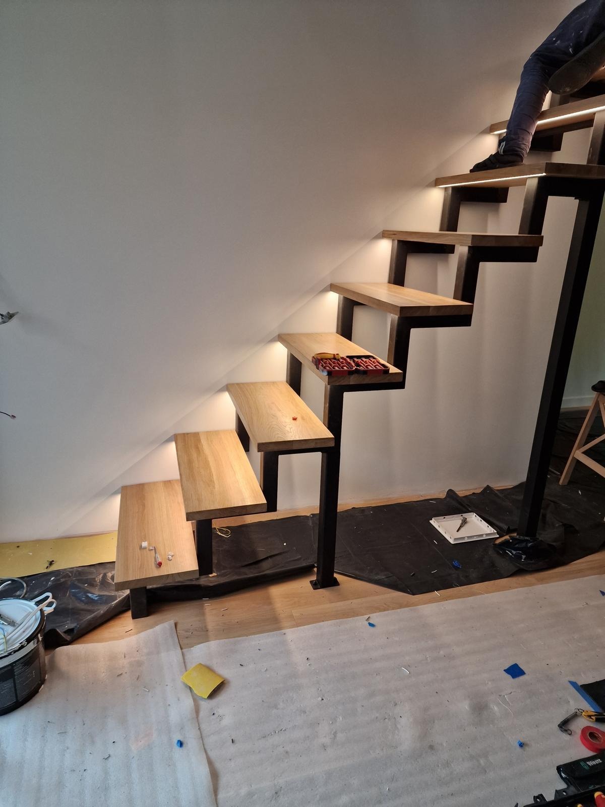 Montaż drewnianych schodów wspornikowych z oświetleniem LED. Widoczne narzędzia i czarny materiał zabezpieczający podłogę. Osoba wchodzi po schodach.