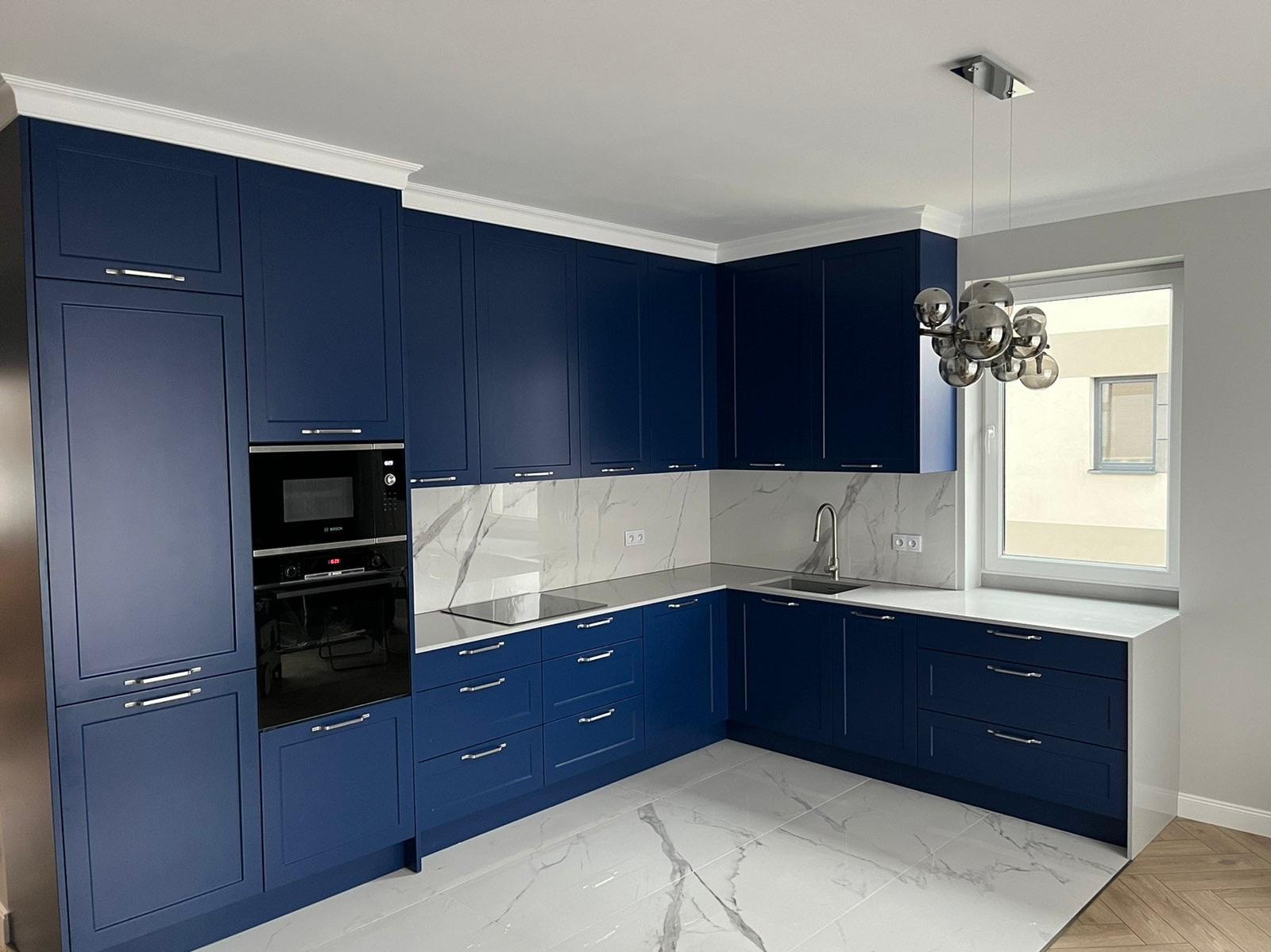 Granatowa zabudowa kuchenna z białymi blatami i marmurowym backsplashiem. Nowoczesne wykończenie wnętrza, srebrny żyrandol i okno z widokiem na zewnątrz.
