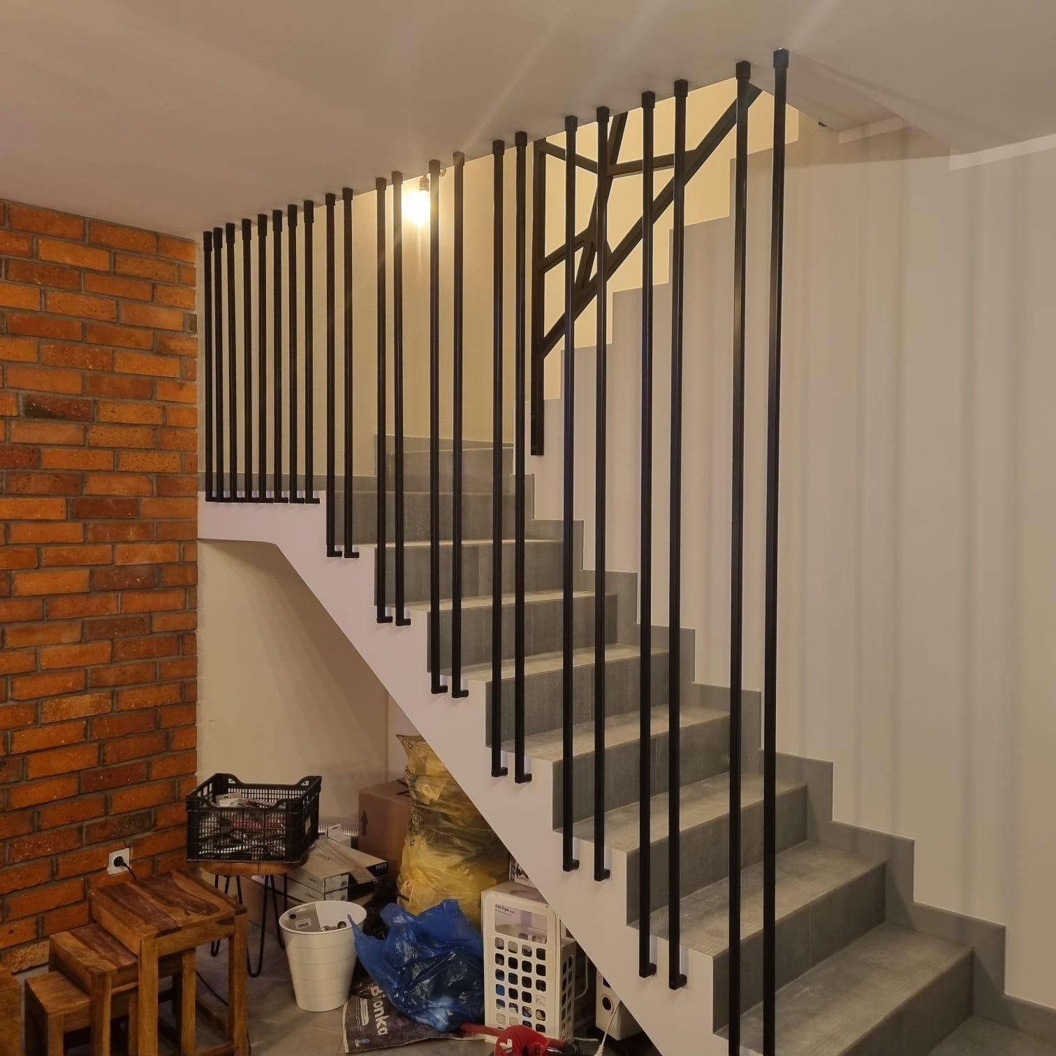 Schody z szarymi stopniami i czarną, minimalistyczną balustradą w nowoczesnym wnętrzu z ceglaną ścianą i widocznymi elementami wykończeniowymi.