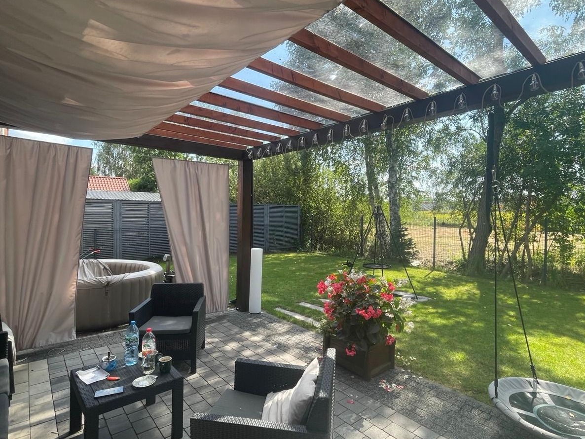 Zadaszona pergola z drewnianą konstrukcją i zasłonami, otoczona zielenią. Na tarasie meble ogrodowe i jacuzzi. Widoczne elementy dekoracyjne i oświetlenie.