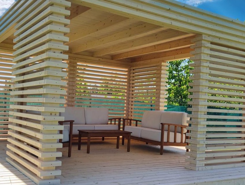 Drewniana pergola z ażurowymi ścianami, wyposażona w sofę i stolik kawowy, tworząca zaciszną strefę relaksu na tarasie. Widok na zieleń w tle.