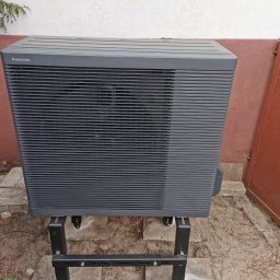 KOVALINSTAL JACEK KOWALIK - Zewnętrzna jednostka klimatyzacji Panasonic, ustawiona na czarnej metalowej podstawie, na tle ściany budynku z elementami zieleni. Widok z bliska, skupiony na urządzeniu.