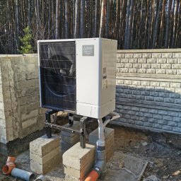 KOVALINSTAL JACEK KOWALIK - Biała pompa ciepła Mitsubishi Electric na zewnątrz, zamontowana na czarnej metalowej konstrukcji, z widocznymi rurami. W tle las i betonowy murek.