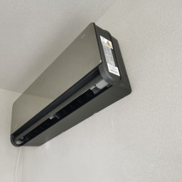 KOVALINSTAL JACEK KOWALIK - Nowoczesny, szary klimatyzator ścienny zamontowany w rogu pokoju na teksturowanej, białej ścianie. Widoczne etykiety informacyjne na obudowie urządzenia.