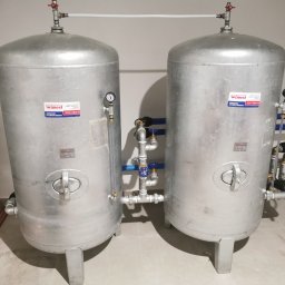 KOVALINSTAL JACEK KOWALIK - Dwa srebrne, pionowe zbiorniki hydroforowe WIMEST ZCS/495/6 z manometrami i rurami, z bliska. Widoczne rury miedziane obok zbiornika i biała ściana w tle.