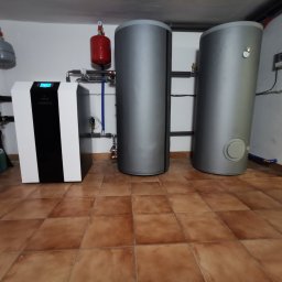 KOVALINSTAL JACEK KOWALIK - Instalacja grzewcza w kotłowni: nowoczesny piec z wyświetlaczem, dwa szare zbiorniki akumulacyjne, instalacja rurowa z czerwonymi zbiornikami wyrównawczymi.