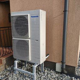 KOVALINSTAL JACEK KOWALIK - Biała, dwuczęściowa pompa ciepła Panasonic na metalowej konstrukcji, zamontowana przy ścianie budynku z rurą spustową. Podłoże wyłożone kamieniami.