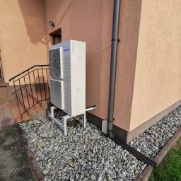 KOVALINSTAL JACEK KOWALIK - Biała, dwuczęściowa pompa ciepła Panasonic, zamontowana na metalowym stelażu przy ścianie budynku z brązową rurą spustową i kamiennym podłożem.