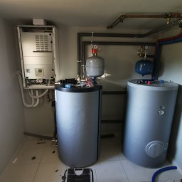 KOVALINSTAL JACEK KOWALIK - Zestaw grzewczy z dwoma zbiornikami akumulacyjnymi, kotłem i orurowaniem w jasnym pomieszczeniu. Widoczne manometry i zawory.