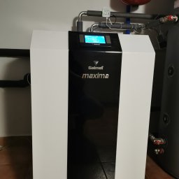 KOVALINSTAL JACEK KOWALIK - Biała pompa ciepła Galmet Maxima z czarnym panelem, widoczny zbiornik buforowy i instalacja rurowa w pomieszczeniu z brązowymi płytkami podłogowymi.