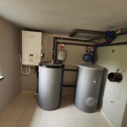 KOVALINSTAL JACEK KOWALIK - Kotłownia z dwoma szarymi zbiornikami akumulacyjnymi, białym piecem gazowym i instalacją rurową. Widok na okno i białe płytki podłogowe.