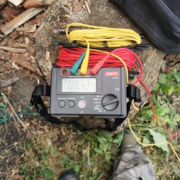 KOVALINSTAL JACEK KOWALIK - Miernik rezystancji uziemienia UNI-T UT521 na pniu, z rozłożonymi kablami pomiarowymi (czerwony, żółty), obok pokrowiec. Widoczne fragmenty butów osoby wykonującej pomiar.