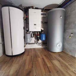 KOVALINSTAL JACEK KOWALIK - Zestaw urządzeń w kotłowni: biały piec gazowy Panasonic, obok dwa cylindryczne zbiorniki (biały i szary), rury i zawory. Podłoga wyłożona panelami imitującymi drewno.