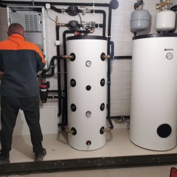 KOVALINSTAL JACEK KOWALIK - Instalacja dwóch białych zbiorników Kospel w kotłowni z czarnymi rurami i armaturą. Pracownik w kurtce montuje instalację. Widoczne płytki na ścianach.