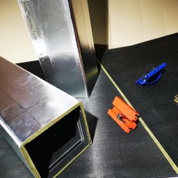 KOVALINSTAL JACEK KOWALIK - Izolowane kanały wentylacyjne o przekroju kwadratowym, folia aluminiowa, zszywacz i klamry na czarnym podłożu. Detal budowy instalacji wentylacyjnej.