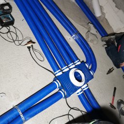 KOVALINSTAL JACEK KOWALIK - Niebieskie rury wentylacyjne, połączone w węzeł z białymi akcentami, tworzą rozbudowany system w surowym wnętrzu. Widoczne narzędzia i elementy montażowe.